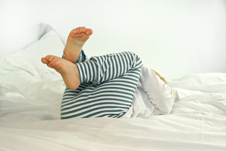 May include: A child wearing green and white striped pajamas lies on a white bed with their feet in the air.