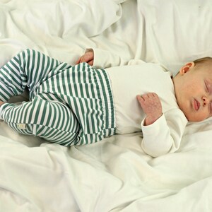 May include: A baby wearing a white long-sleeved onesie and green and white striped pants is sleeping on a white sheet.