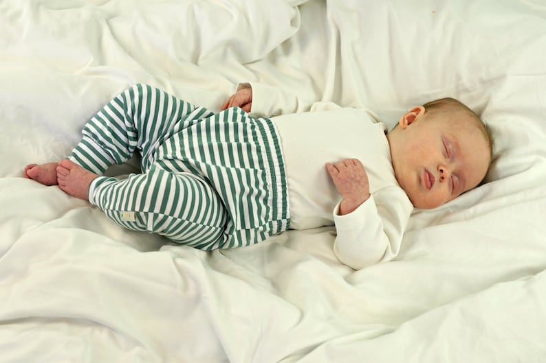 May include: A baby wearing a white long-sleeved onesie and green and white striped pants is sleeping on a white sheet.
