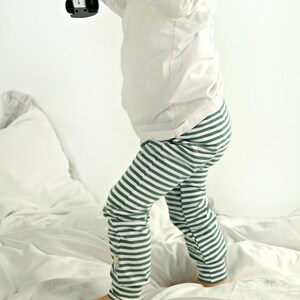 May include: A child wearing a white long-sleeved shirt and green and white striped pants stands on a white bed. The child is holding a blue toy car.