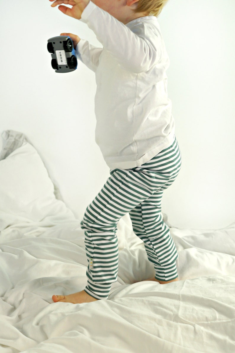 May include: A child wearing a white long-sleeved shirt and green and white striped pants stands on a white bed. The child is holding a blue toy car.