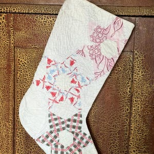 May include: A quilted Christmas stocking with a white background and star patterns. The star patterns are red, blue, and green plaid. The top has a floral pattern and a loop for hanging.