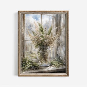 May include: A framed watercolour print depicting a window scene. A glass jar filled with dried flowers and greenery sits on a windowsill. The window reveals a mountain landscape and a cloudy sky. The frame is made of light-coloured wood.