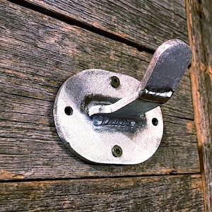 May include: A silver-colored metal wall hook with an oval-shaped base plate. The hook features a prominent welded joint connecting the upright arm to the base. It is secured to a wooden surface with two visible dark-colored screws at the top and bottom.
