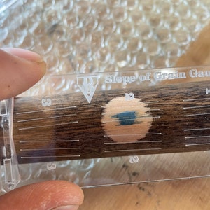 May include: A clear plastic Slope of Grain Gauge with a black diamond logo and the text "Slope of Grain Gauge". The gauge has a numbered scale from 1 to 3. A dark brown wood surface is visible through the gauge.