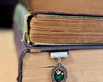 May include: A stack of antique books with aged, tan pages. A decorative silver pendant with a floral design hangs from a ribbon bookmark. The pendant features white flowers and green leaves on a black background.