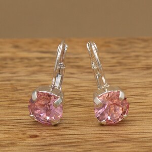May include: A pair of leverback earrings featuring round, faceted pink stones set in silver-toned metal prongs. The earrings have a classic design with a secure hinged closure mechanism, showcasing the clear, light pink color of the stones.