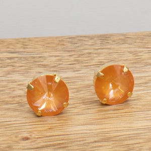 May include: Two orange crystal stud earrings with gold-colored settings. The earrings are round and faceted, reflecting light. The earrings are set on a wooden surface.