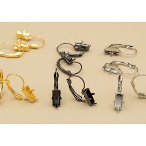 May include: Three pairs of earring settings, one pair in gold, one pair in black, and one pair in silver. Each setting has a rectangular bezel for a stone or cabochon.