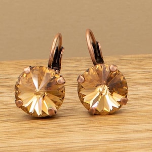 May include: A pair of leverback earrings featuring large, round, faceted crystals in a warm amber color. The earrings have a copper-colored metal setting and leverback closures, resting on a wooden surface.