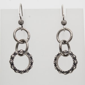 May include: A pair of silver earrings with a unique design. Each earring features a small, textured ball at the top, connected to two interlocking rings. The bottom ring is adorned with a floral pattern, creating a delicate and stylish accessory.