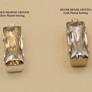 May include: Two rectangular crystal pendants. The left pendant is a Golden Shadow crystal in a silver-plated setting. The right pendant is a Silver Shade crystal in a gold-plated setting. Each pendant features a small metal loop at the top.