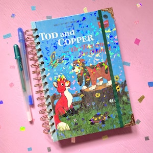 Peut inclure: Un carnet à spirales avec une couverture holographique portant le texte TOD and COPPER et The Fox and the Hound. La couverture montre un renard, un chien de chasse, un papillon et une tortue dans un champ. Deux stylos bleus sont posés à côté.