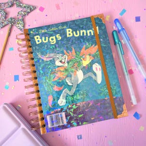 May include: A spiral-bound notebook featuring a holographic cover with an illustration of Bugs Bunny holding carrots. The cover displays the text 'a Little Golden Book Bugs Bunny' and includes a barcode, a copper-coloured metal spiral binding, and a matching elastic closure band.