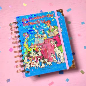 May include: A spiral-bound notebook featuring a One Hundred and One Dalmatians illustration on the cover. The blue cover displays characters playing a piano, surrounded by puppies, with a holographic finish, copper-coloured wire binding, and an elastic band.