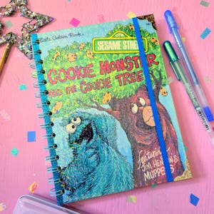 May include: A spiral-bound notebook featuring a Little Golden Book cover titled Sesame Street Cookie Monster and the Cookie Tree. The cover displays a blue Cookie Monster and a brown bear, with a vertical blue elastic band and gold corner accents.