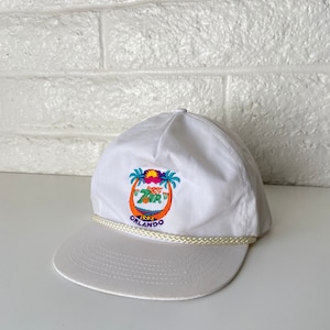 May include: White baseball cap with a white rope trim. The front of the cap features an embroidered logo with palm trees, a sunset, and the text "1993 ORLANDO".