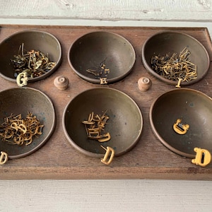 May include: A wooden tray with six brass bowls, each containing small metal objects. The bowls are arranged in two rows of three. Gold-colored letters are placed near the bowls. The tray sits on a white wooden surface.
