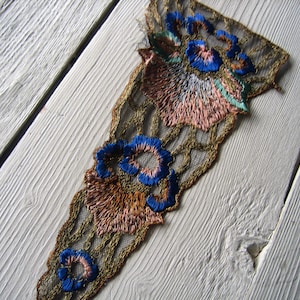 May include: A gold lace trim with embroidered flowers in shades of blue, pink, and brown. The trim is on a white wooden surface.
