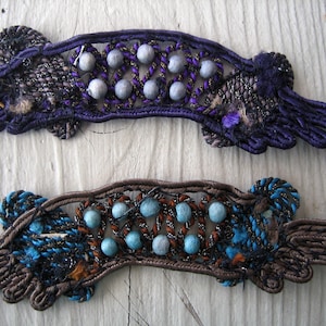 May include: Two decorative fabric trims with a woven design and blue and silver beads. The trim on top is purple and the trim on the bottom is brown.