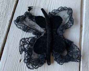 May include: A black butterfly-shaped decorative item. The wings are made of black lace with a scalloped edge, and the body is a solid black material. The butterfly has a wire antenna and is set against a white wooden surface.