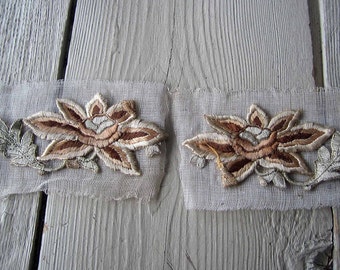 May include: Two beige and brown embroidered floral appliques on a white mesh fabric. The appliques are in the shape of flowers with leaves.