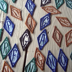 May include: A collection of diamond-shaped fabric patches with letters embroidered on them. The patches are in various colors, including blue, green, brown, and black. The letters are in a variety of fonts and styles.