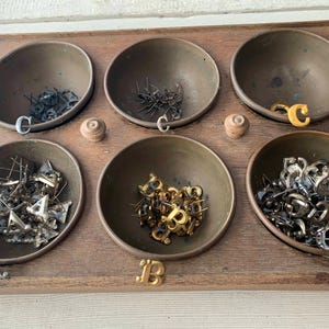 May include: A wooden box with six brass bowls filled with various metal and gold letter charms. The bowls are labeled with the letters A, B, and C.