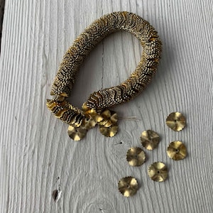 May include: A vintage-style hair accessory featuring a looped, textured band and a cluster of decorative flower-shaped elements. Additional loose, flower-shaped pieces are scattered nearby. The piece is made of gold-toned metal and rests on a white wooden surface.