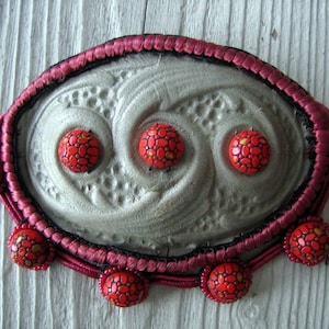 May include: A red and black beaded necklace with a large, gray, oval-shaped pendant featuring a swirling design. The pendant is surrounded by a red cord and black thread.