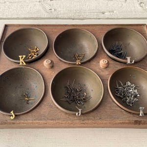May include: A wooden tray with six small brass bowls, each filled with different types of nails and tacks. The bowls are labeled with the letters J, K, and L.