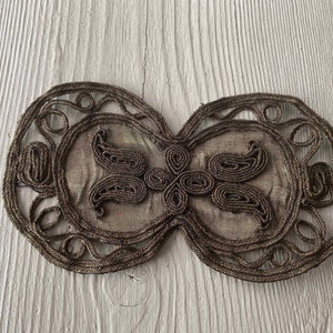 May include: A decorative, gold-colored, embroidered fabric embellishment with a paisley design. The embellishment is in the shape of a butterfly and has a delicate, intricate pattern.