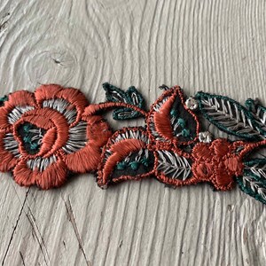 May include: A close-up of a floral embroidered patch with red, white, and green thread. The patch features two flowers and leaves.