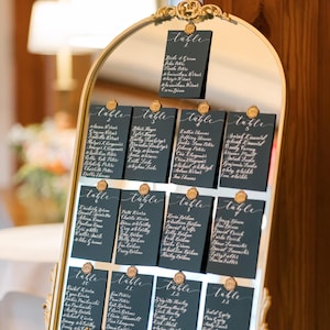 May include: A gold-framed mirror displaying black table cards with white calligraphy. Each card features guest names and table numbers, sealed with a gold wax stamp. The arrangement is set on a gold easel, ideal for a wedding or event.