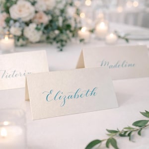 May include: Ivory place cards with elegant teal calligraphy, featuring names like "Elizabeth" and "Madeline." The cards are set on a white tablecloth, with candles, flowers, and greenery in the background, creating a soft, romantic ambiance.