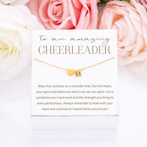 May include: A white card with gold lettering that reads "to an amazing CHEERLEADER". The card is next to a gold necklace with a heart charm and a letter "M" charm. The card reads "Wear this necklace as a reminder that, like this heart, your spirit and talent are what truly set you apart. Let it symbolize your hard work and the strength you bring to every performance. Always remember to lead with your heart and continue to inspire those around you!"