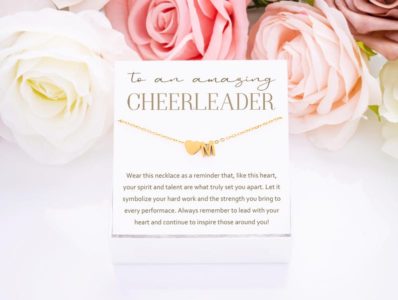 May include: A white card with gold lettering that reads "to an amazing CHEERLEADER". The card is next to a gold necklace with a heart charm and a letter "M" charm. The card reads "Wear this necklace as a reminder that, like this heart, your spirit and talent are what truly set you apart. Let it symbolize your hard work and the strength you bring to every performance. Always remember to lead with your heart and continue to inspire those around you!"