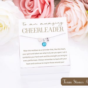 May include: A white card with the text "to an amazing CHEERLEADER" and a silver necklace with a heart charm, a letter charm, and a blue gemstone charm. The card also includes a message about the necklace symbolizing hard work and strength. The text "Team Stones Available!" is at the bottom of the card.