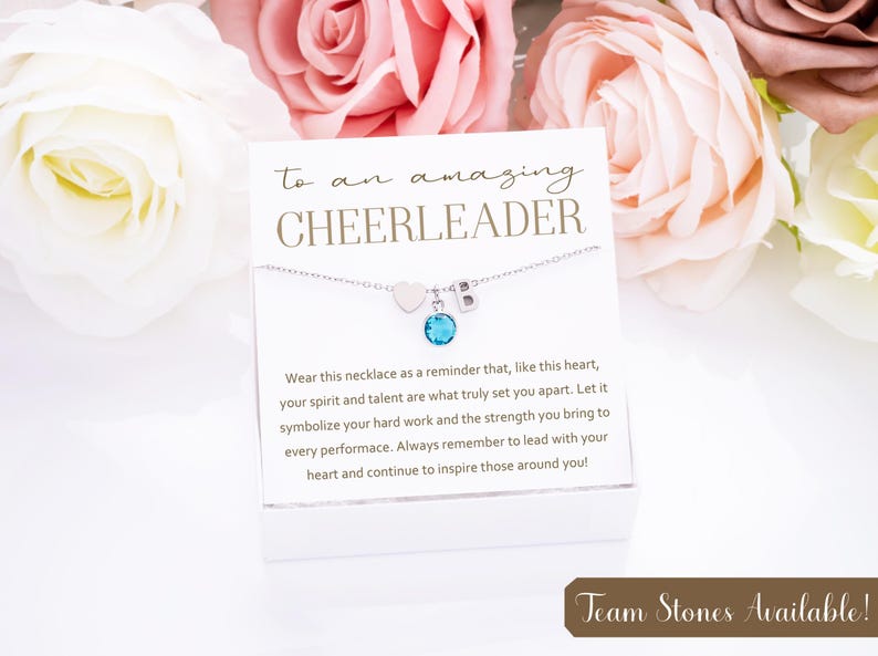 May include: A white card with the text "to an amazing CHEERLEADER" and a silver necklace with a heart charm, a letter charm, and a blue gemstone charm. The card also includes a message about the necklace symbolizing hard work and strength. The text "Team Stones Available!" is at the bottom of the card.