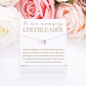 May include: A white card with the text "to an amazing CHEERLEADER" and a silver necklace with a heart and letter charm. The card has a message about the necklace being a reminder to lead with your heart and inspire those around you.