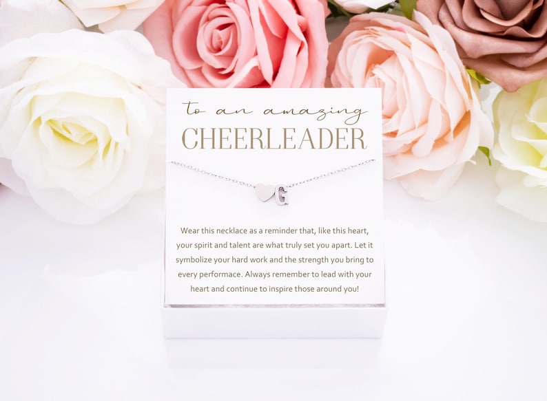 May include: A white card with the text "to an amazing CHEERLEADER" and a silver necklace with a heart and letter charm. The card has a message about the necklace being a reminder to lead with your heart and inspire those around you.