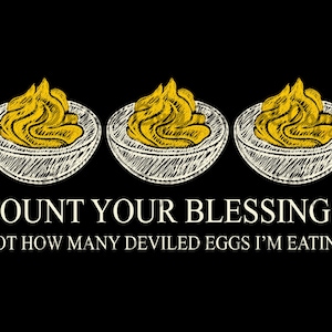 May include: A black graphic featuring three white sketched deviled eggs with yellow yolks. Below the eggs, white text reads: COUNT YOUR BLESSINGS, NOT HOW MANY DEVILED EGGS I'M EATING. The design uses a hand-drawn, cross-hatched style for the food.