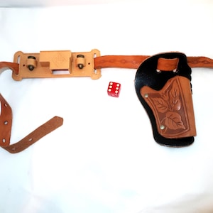 May include: A brown leather toy gun holster and belt set with a decorative leaf pattern on the holster. The set includes a red die. The belt has a buckle and a decorative pattern.