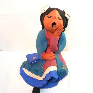 May include: A ceramic figurine of a person yawning, with closed eyes and mouth open. The figure wears a blue and pink dress, a blue shawl, and has black hair with white accents. A blue book with a gold star is on the lap.