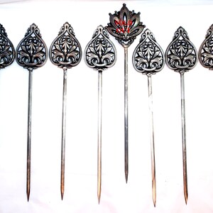 May include: A collection of silver-toned cocktail picks with ornate, teardrop-shaped tops featuring intricate floral designs. One pick has a crown-like top with red accents. The picks have long, slender stems and are arranged on a white surface.