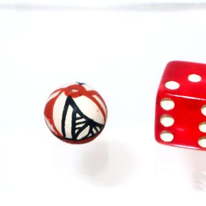 May include: A small, round, off-white bead featuring hand-painted red and black geometric line patterns, positioned next to a translucent red six-sided die with white pips on its faces, providing a clear visual scale for the bead's size.