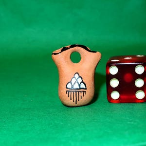 May include: A small, brown, ceramic figurine with a hole and a painted design of a triangle and lines, next to a red die with white dots. The background is a solid green.