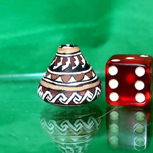 May include: A small, hand-painted ceramic vase with a brown, black, and white geometric design, next to a red, translucent die with white dots. The vase is approximately 5 cm tall.