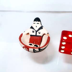 May include: A small, hand-painted ceramic figurine of a person sitting inside a bowl-shaped vessel. The figure has black hair, a white shirt with black trim, and red pants. The bowl is cream-colored with red and black painted accents.