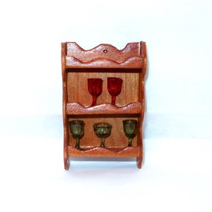 May include: A miniature wooden wine rack with three shelves, holding two red wine glasses on the top shelf and three green glasses on the bottom shelf. The rack is made of stained wood and has a decorative scalloped top.